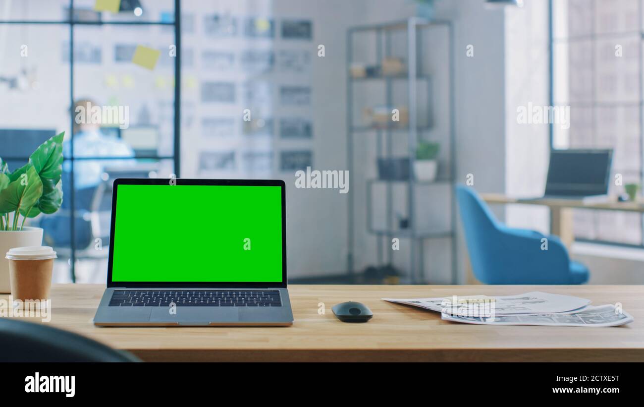 Laptop on the Desk in the Office Shows Green Mock-up Screen. In the Background Creative Office or Business HUB with Professional Working Stock Photo