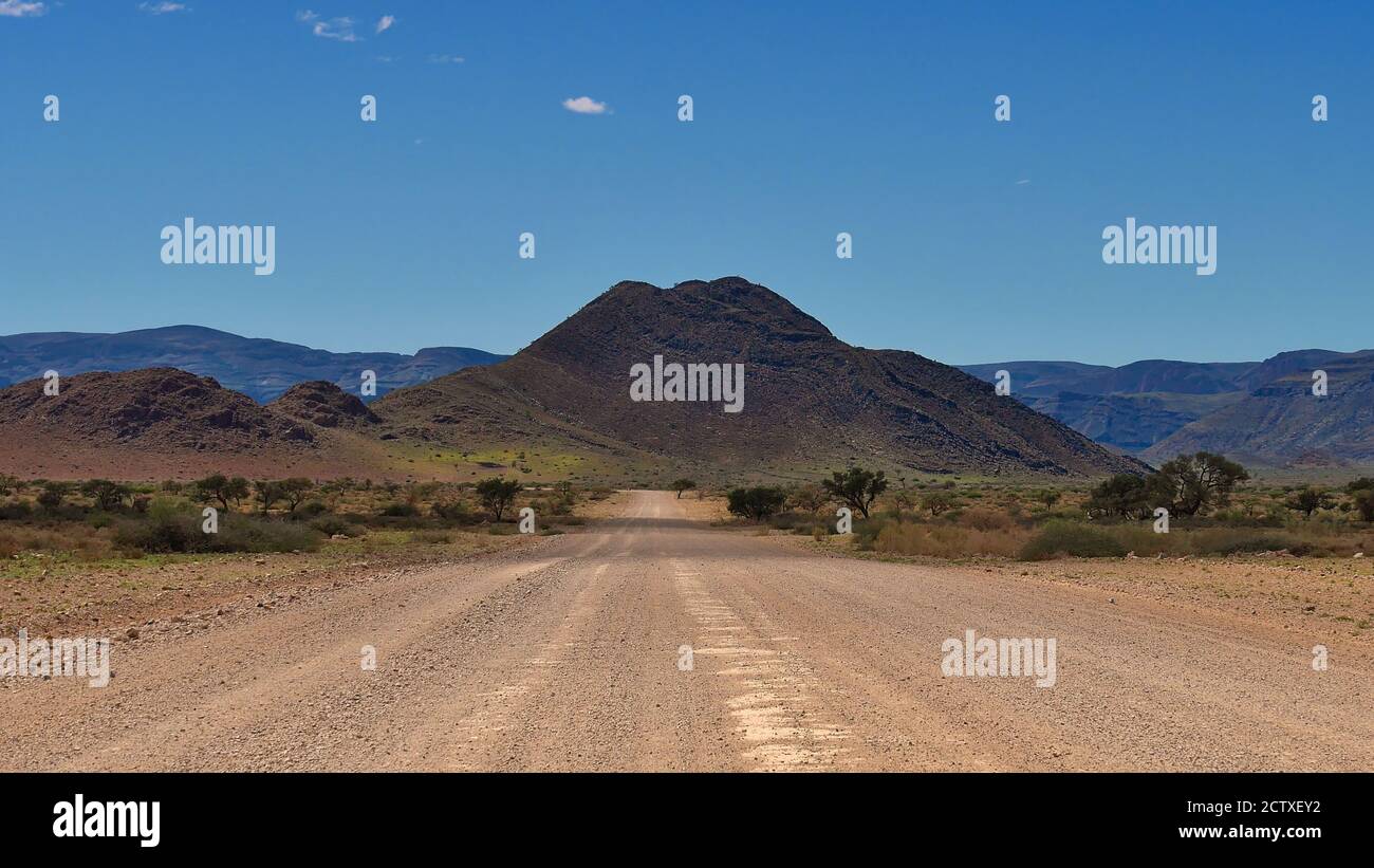 Gravel road leading towards a mountain with slightly green grass and some trees shortly after the end of rainy season, Sesriem, Namib desert, Namibia. Stock Photo