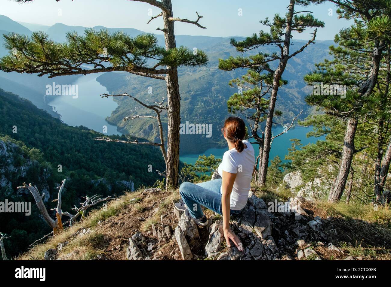 Young female nature explorer enjoying the panoramic view on a canyon of river Drina from Tara mountain viewpoint, sitting on a stone cliff. Hiking thr Stock Photo