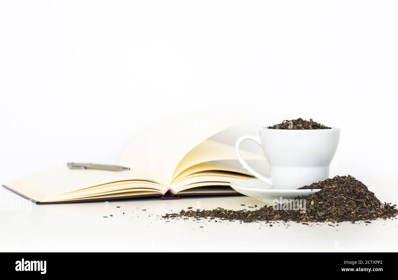 Dry tea leaves overflowing from a cup next to an open notebook and a pen isolated against a white background. Stock Photo