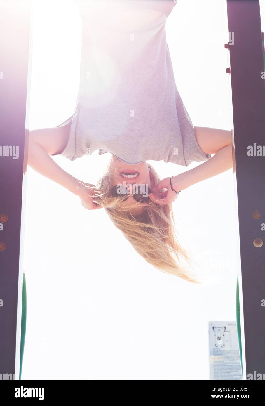 Happy female model hanging upside down at the playground wearing sunglasses. Stock Photo