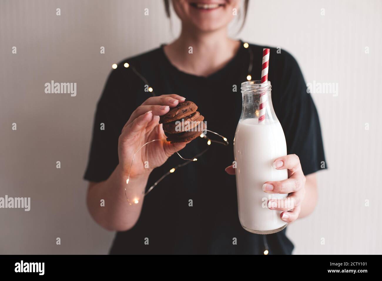 Smiling woman eating chocolate cookies and holding bottle of milk in room close up. Winter holiday season. Good morning. Stock Photo