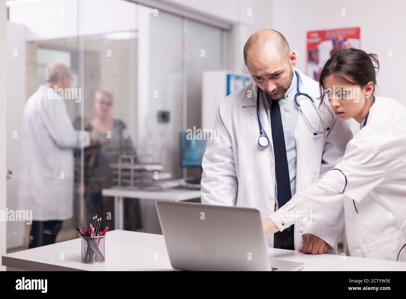 Team of young doctors working together in hospital checking patient evolution on laptop. Elderly aged woman and senior medic on clinic corridor. Physician wearing white coat and stethoscope. Stock Photo
