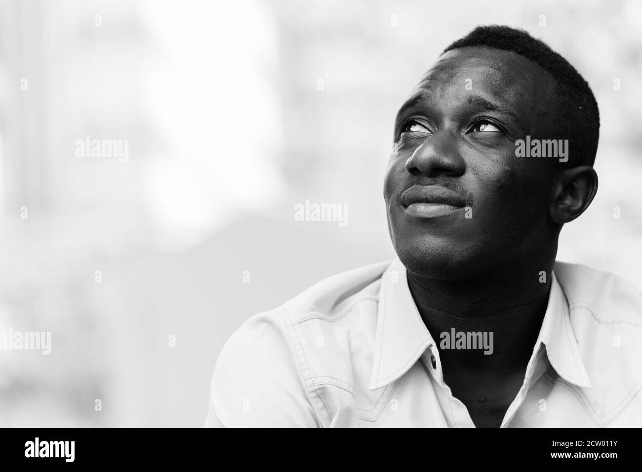 Young black African man sitting while thinking and looking up outside coffee shop against view of nature and city Stock Photo