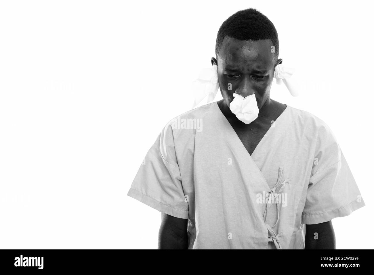 Studio shot of young black African man patient looking down and sick with tissues in his ears and nose Stock Photo