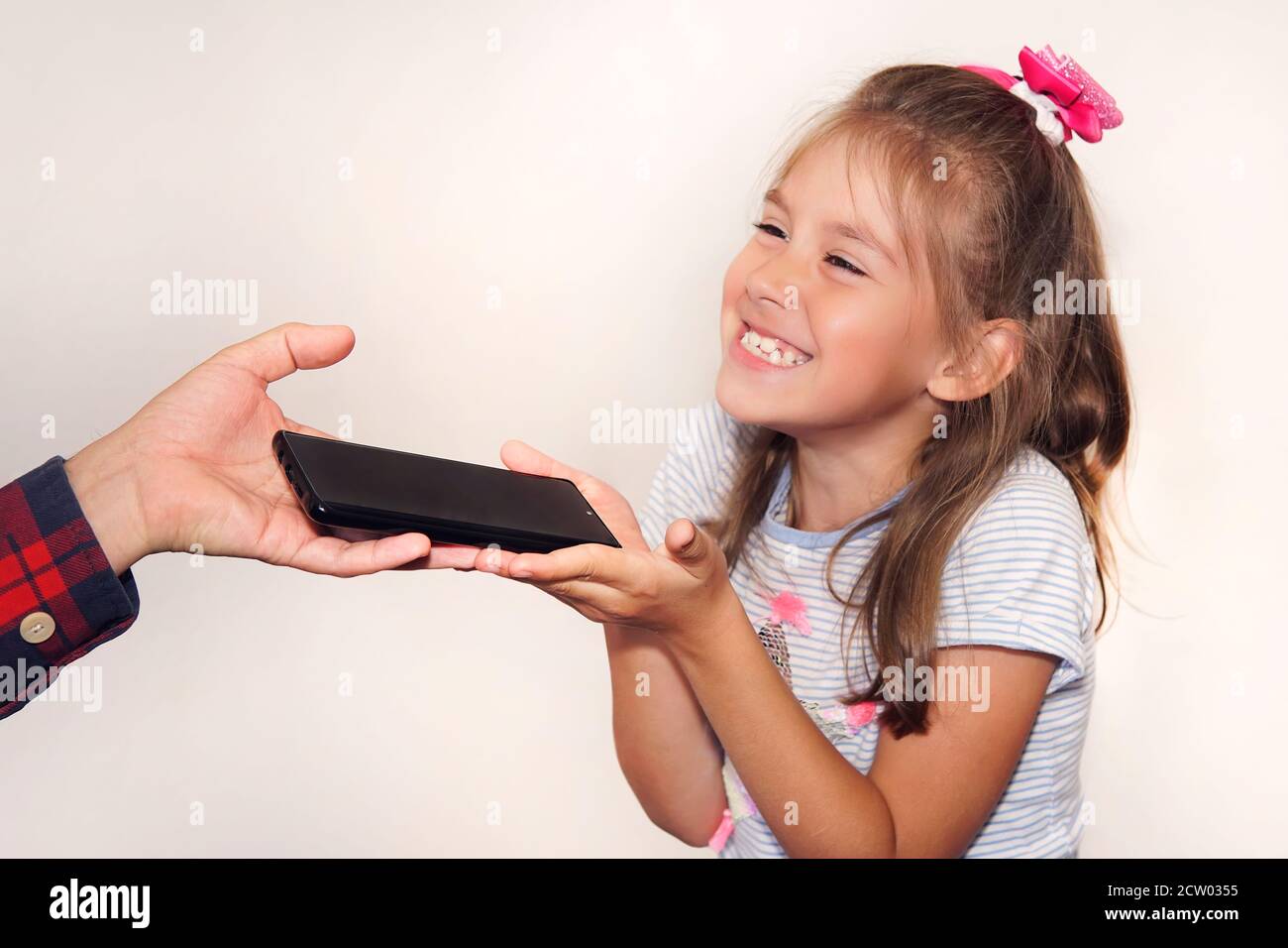 father gives a gift to his daughter on her birthday. A man's hand passes a black new smartphone to the girl in close-up. A happy child grabs the phone Stock Photo