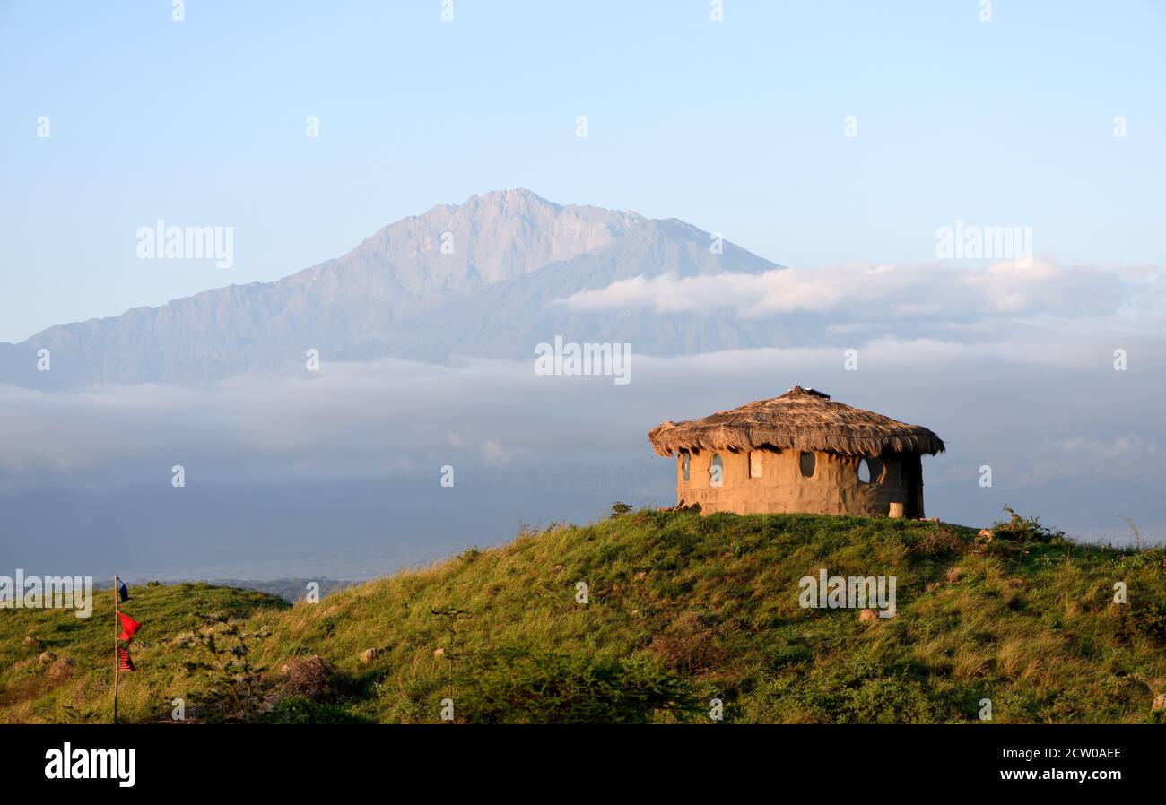 A round, thatched, earth walled building with the south-eastern slopes of the dormant volcano Mount Meru in the background.  Sanya Juu, Boma Ngombe, T Stock Photo