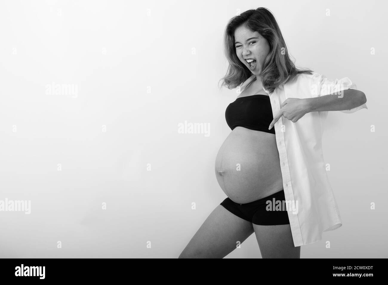 Studio shot of young happy Asian pregnant woman smiling while pointing at her stomach and winking Stock Photo