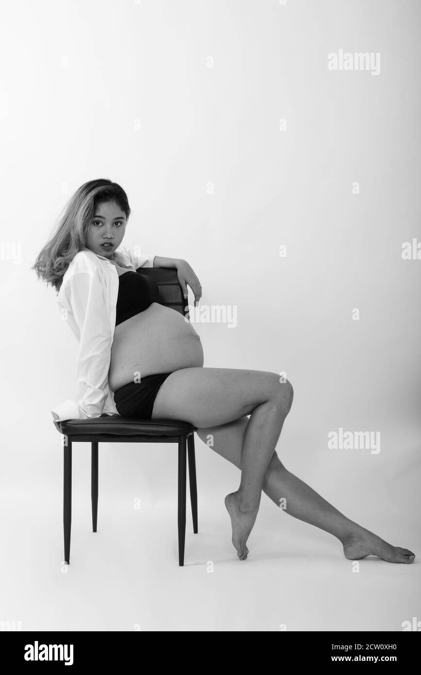 Full body shot of young Asian pregnant woman posing while sitting on chair against white background Stock Photo