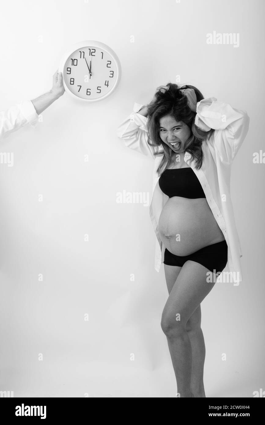 Studio shot of young Asian pregnant woman looking stressed with countdown on wall clock against white background Stock Photo