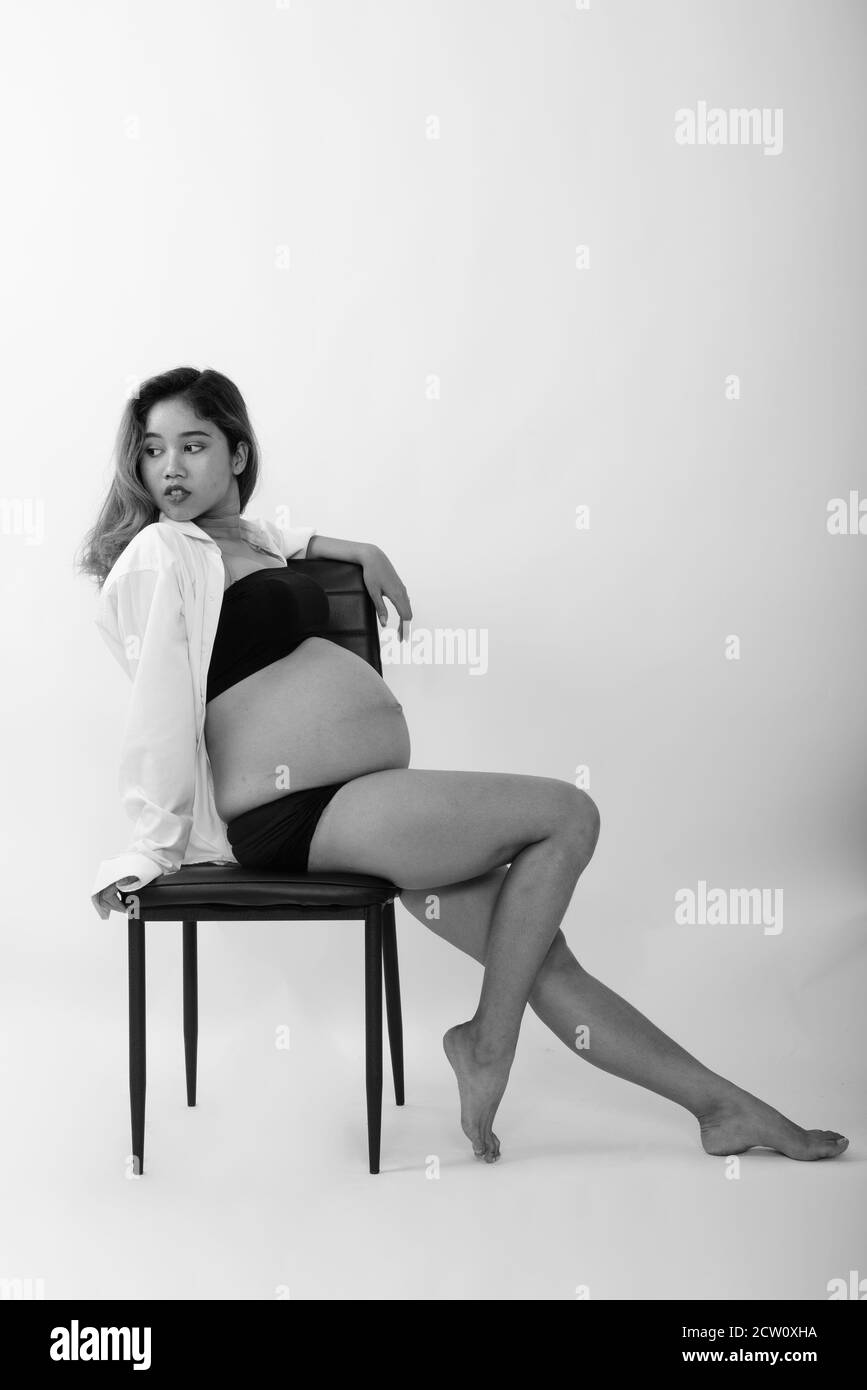 Full body shot of young Asian pregnant woman thinking and posing while sitting on chair against white background Stock Photo