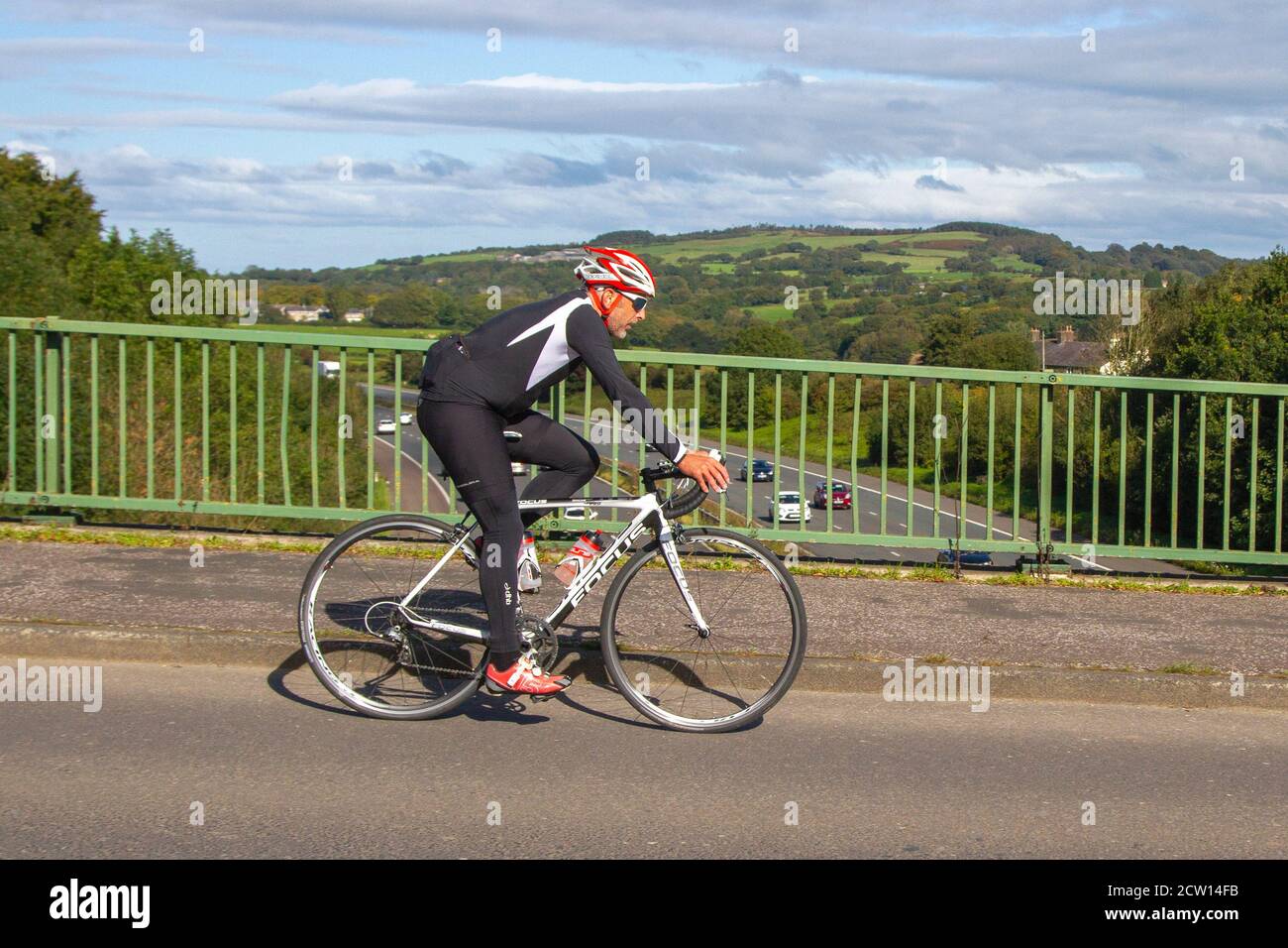 Male cyclist wearing spiuk helmet riding white FOCUS Izalco Pro 3.0 Compact on countryside route crossing motorway bridge in rural Lancashire, UK Stock Photo