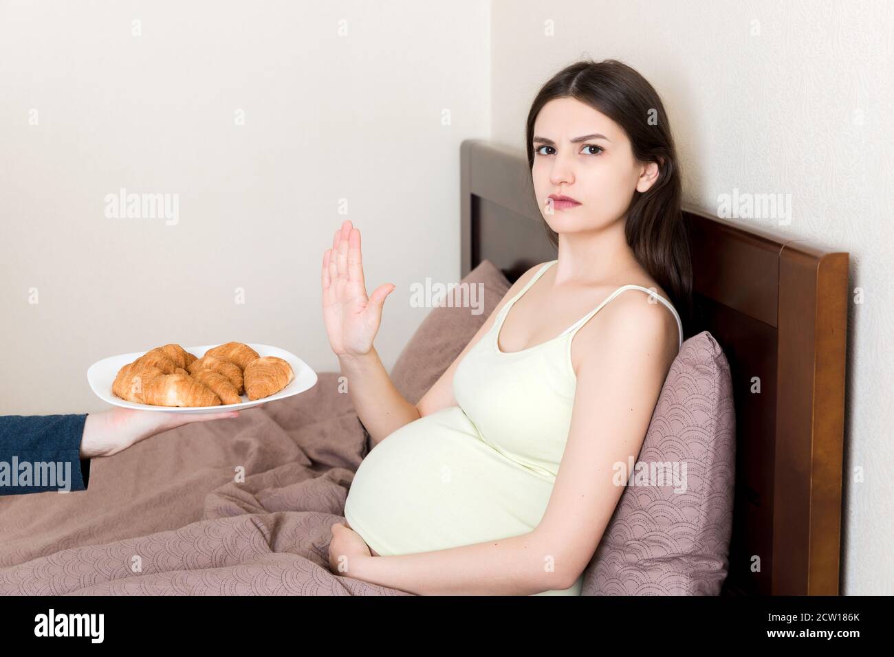 Pregnant woman relaxing in bed makes stop gesture to croissants