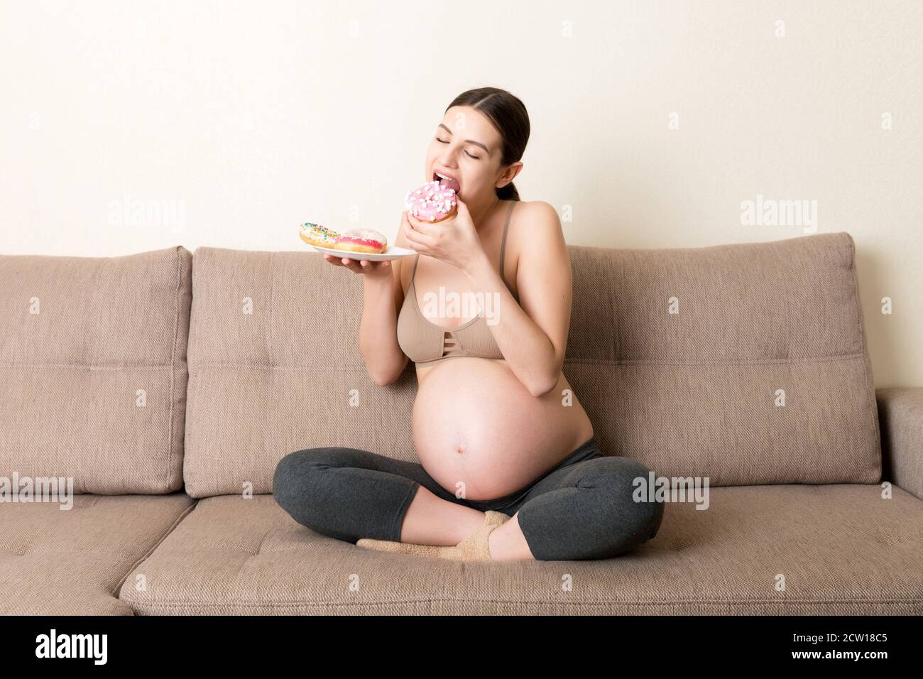 Pregnant woman is eating many donuts relaxing on the sofa. Unhealthy dieting during pregnancy concept. Stock Photo