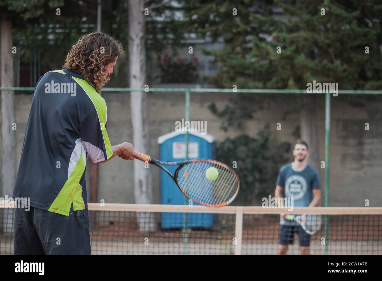 Man with longer hair seen from behind swining his racquet forehand to hit the ball, playing tennis outdoors with a friend. Ball seen blurred flying fa Stock Photo