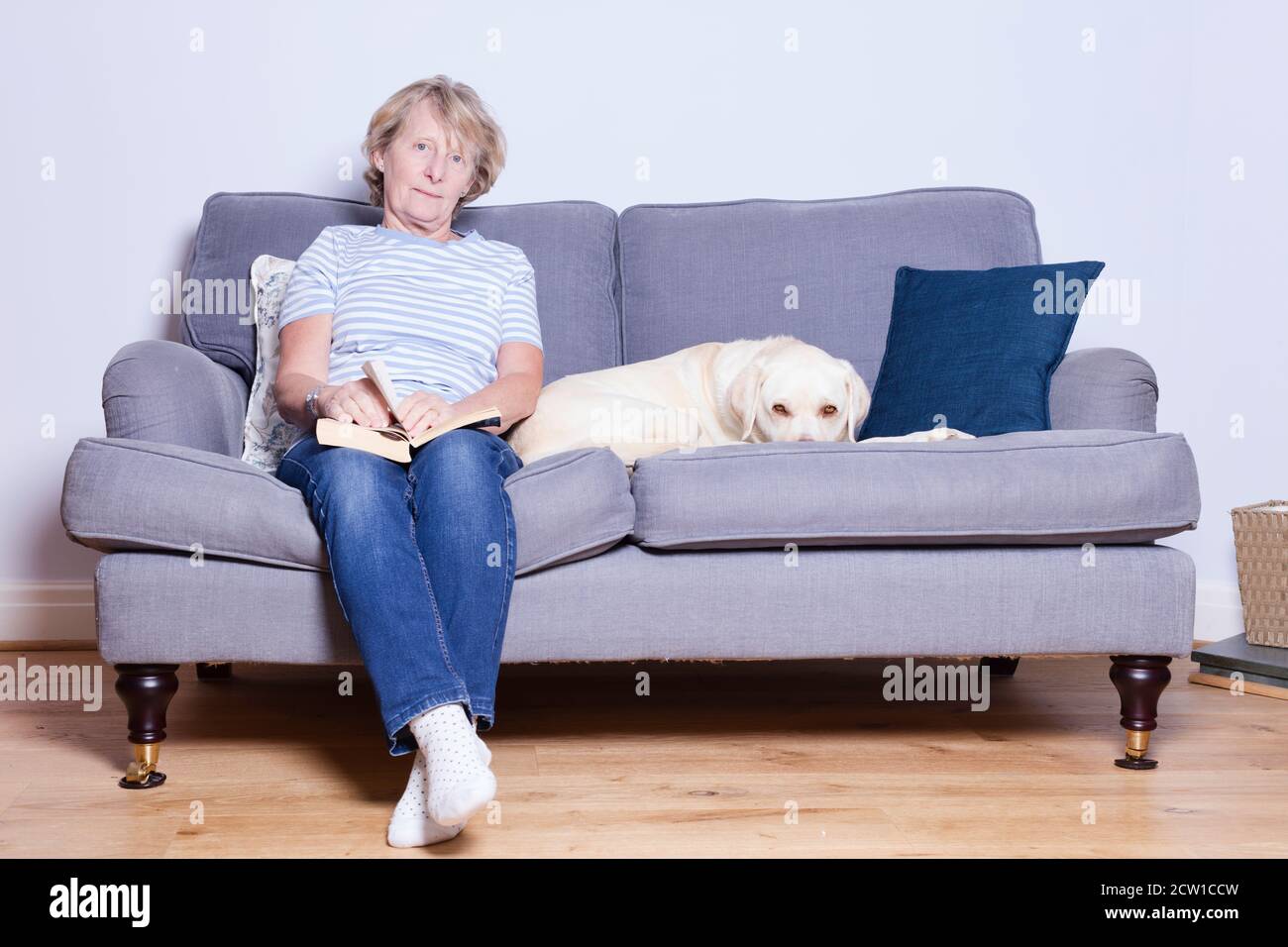 Lady in her late 60s relaxing on the sofa with a book and her dog Stock ...