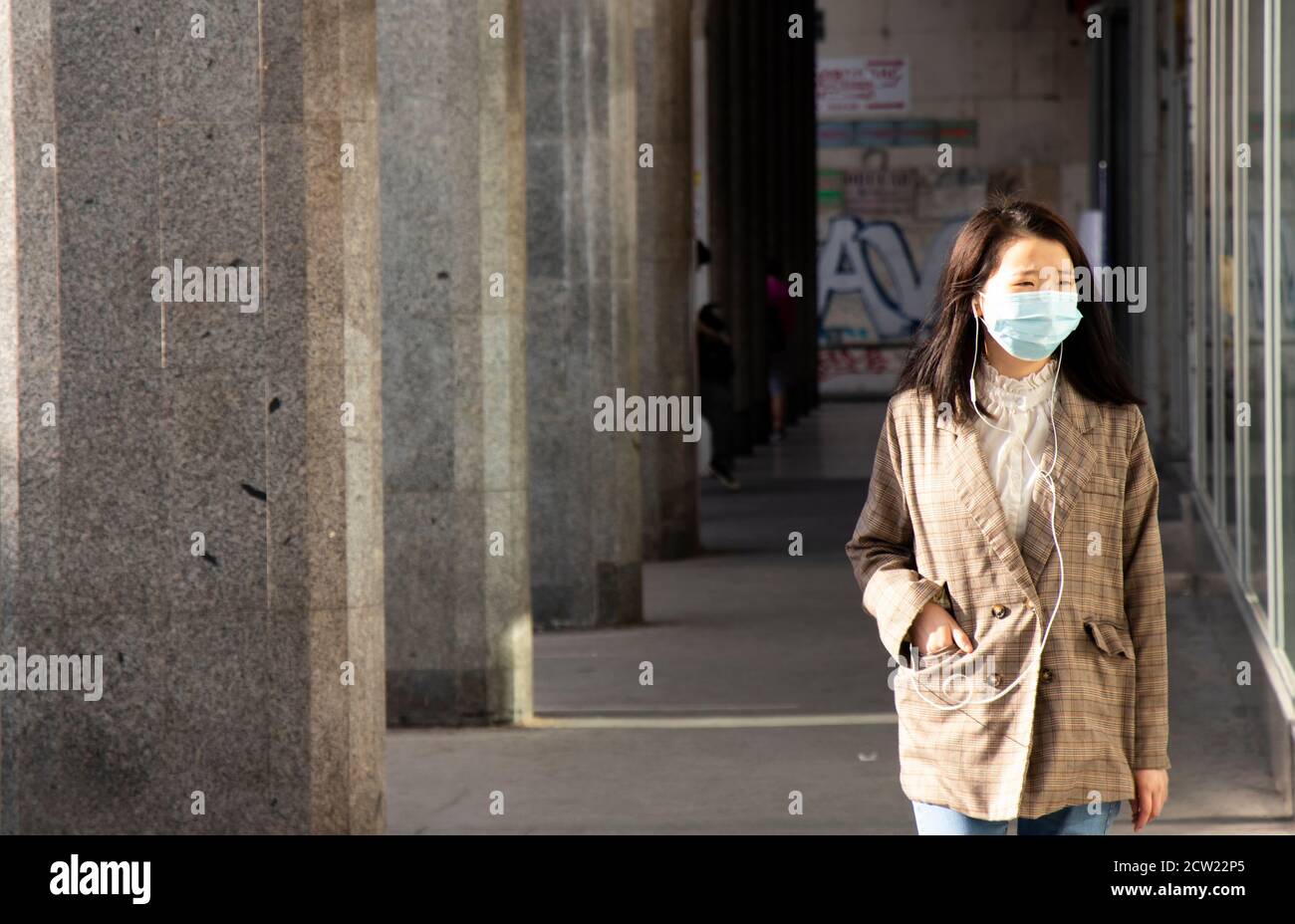 Belgrade, Serbia - September 26, 2020: Young Asian women wearing face surgical mask and earphones walking down the street with a hand in a pocket Stock Photo