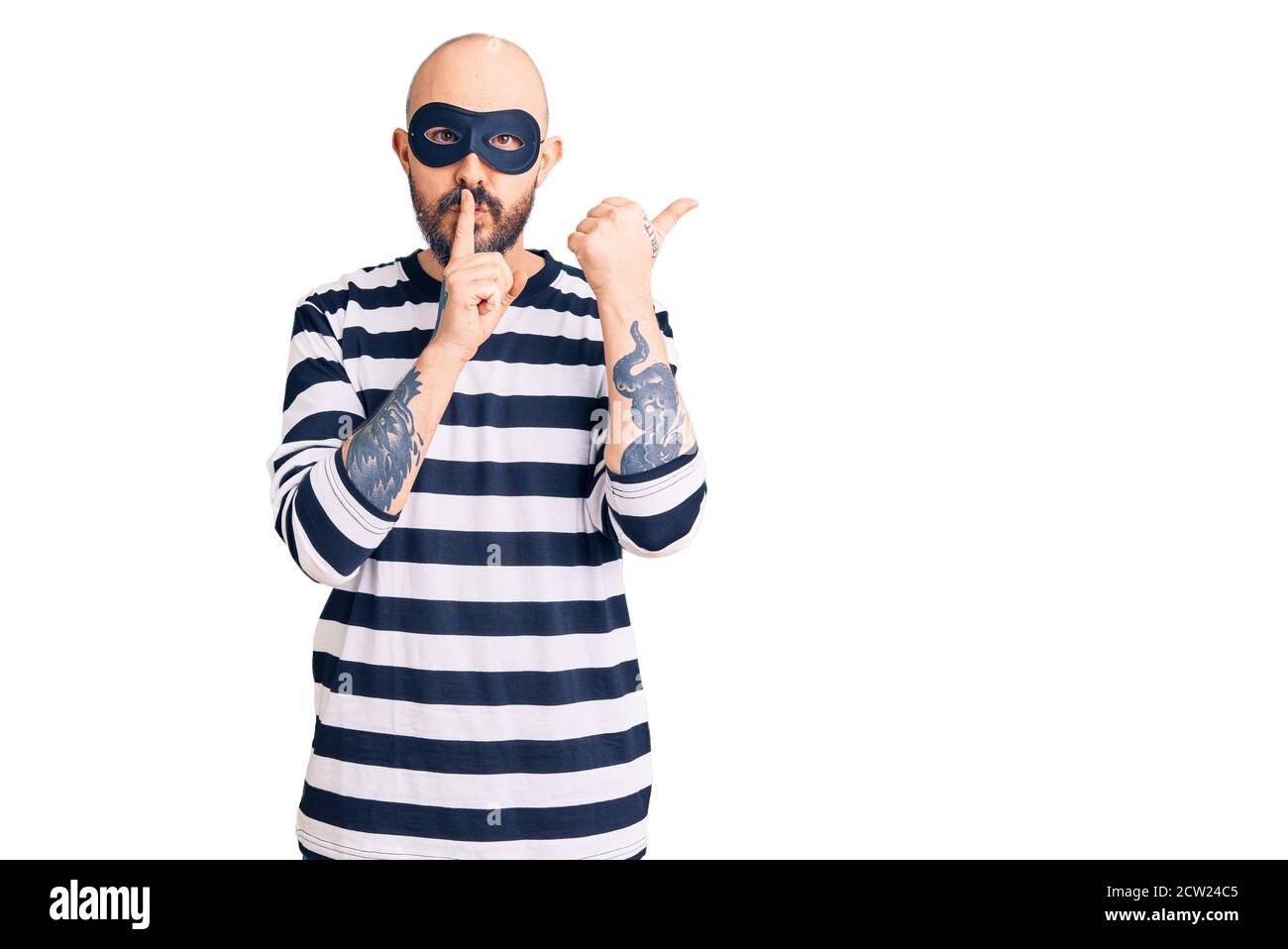 Young handsome man wearing burglar mask asking to be quiet with finger on lips pointing with hand to the side. silence and secret concept. Stock Photo