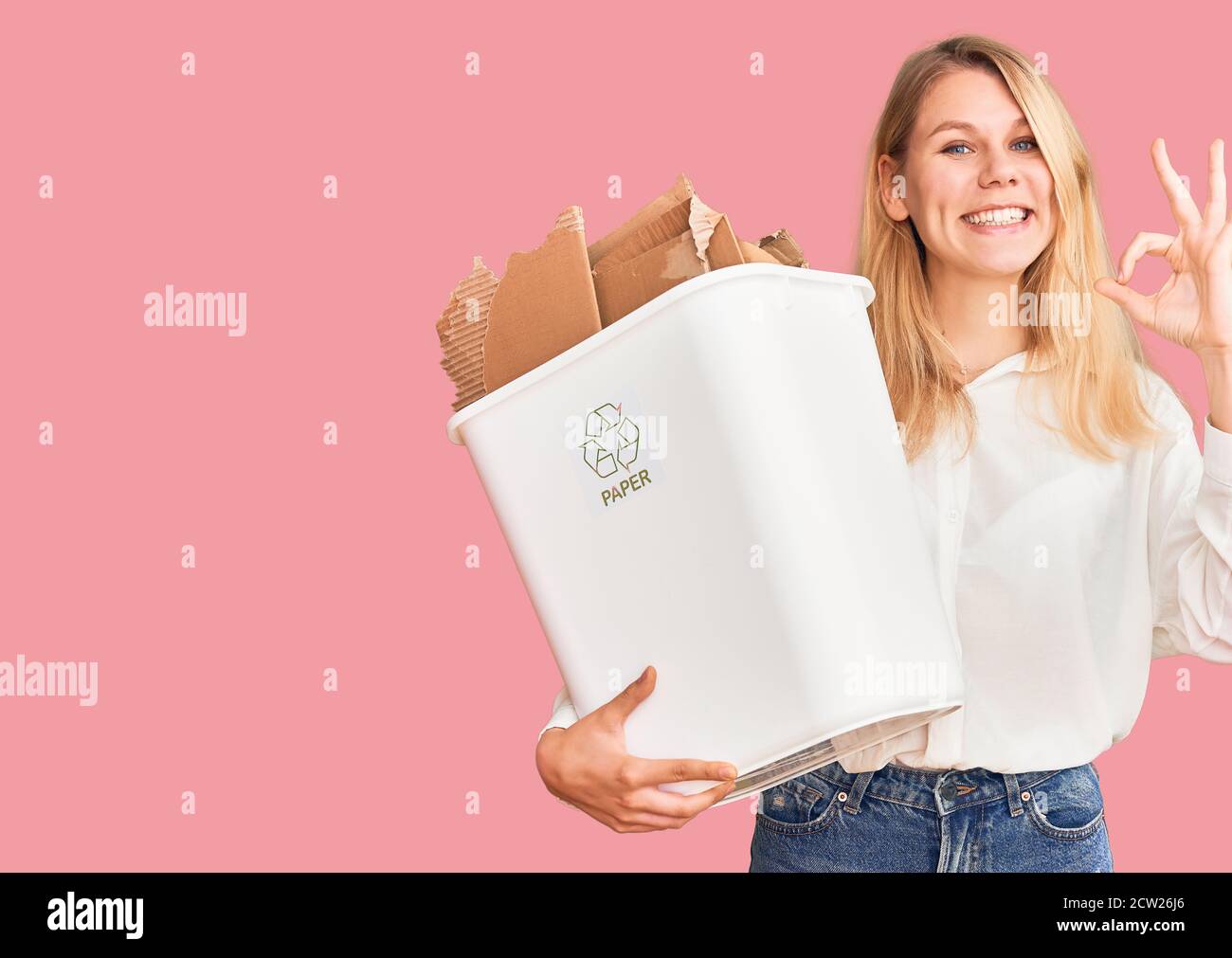 Young beautiful blonde woman holding recycle paper bin doing ok sign with fingers, smiling friendly gesturing excellent symbol Stock Photo