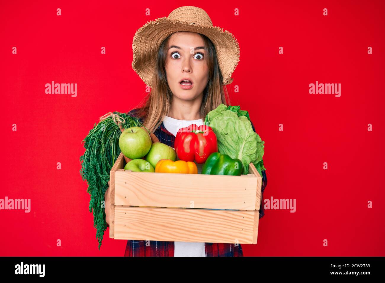 Beautiful caucasian woman wearing farmer hat holding vegetables afraid ...