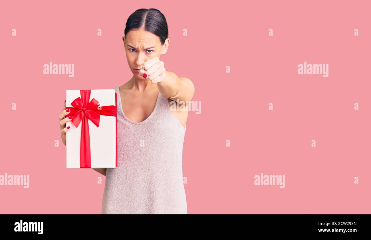Beautiful brunette young woman holding gift annoyed and frustrated shouting with anger, yelling crazy with anger and hand raised Stock Photo