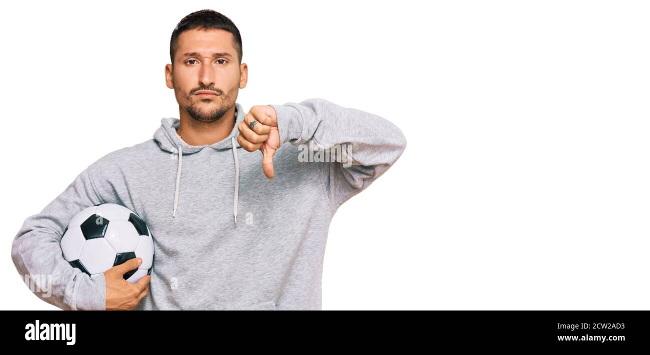 Handsome man with tattoos holding soccer ball with angry face, negative sign showing dislike with thumbs down, rejection concept Stock Photo