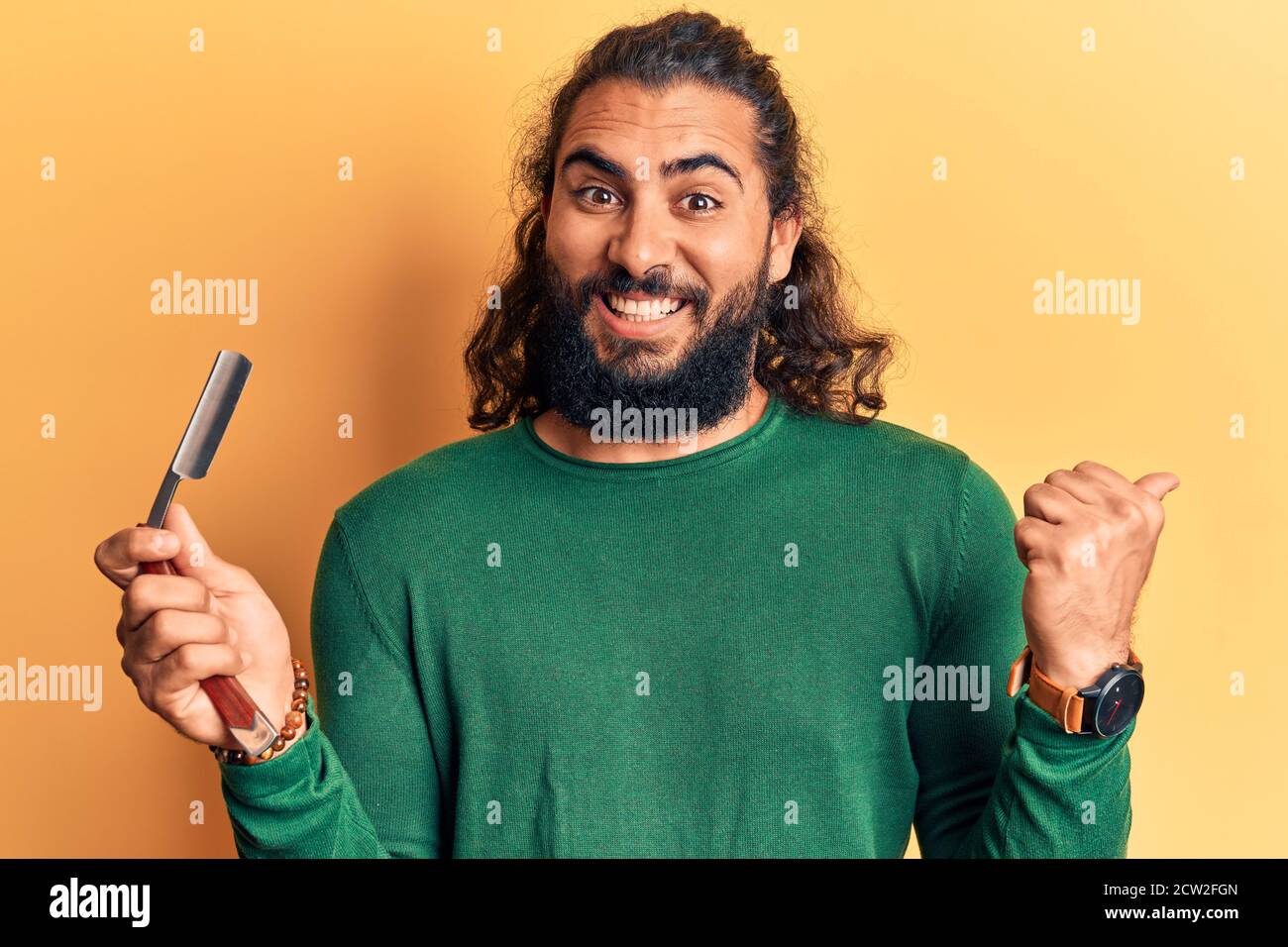 Young arab man holding barber razor pointing thumb up to the side
