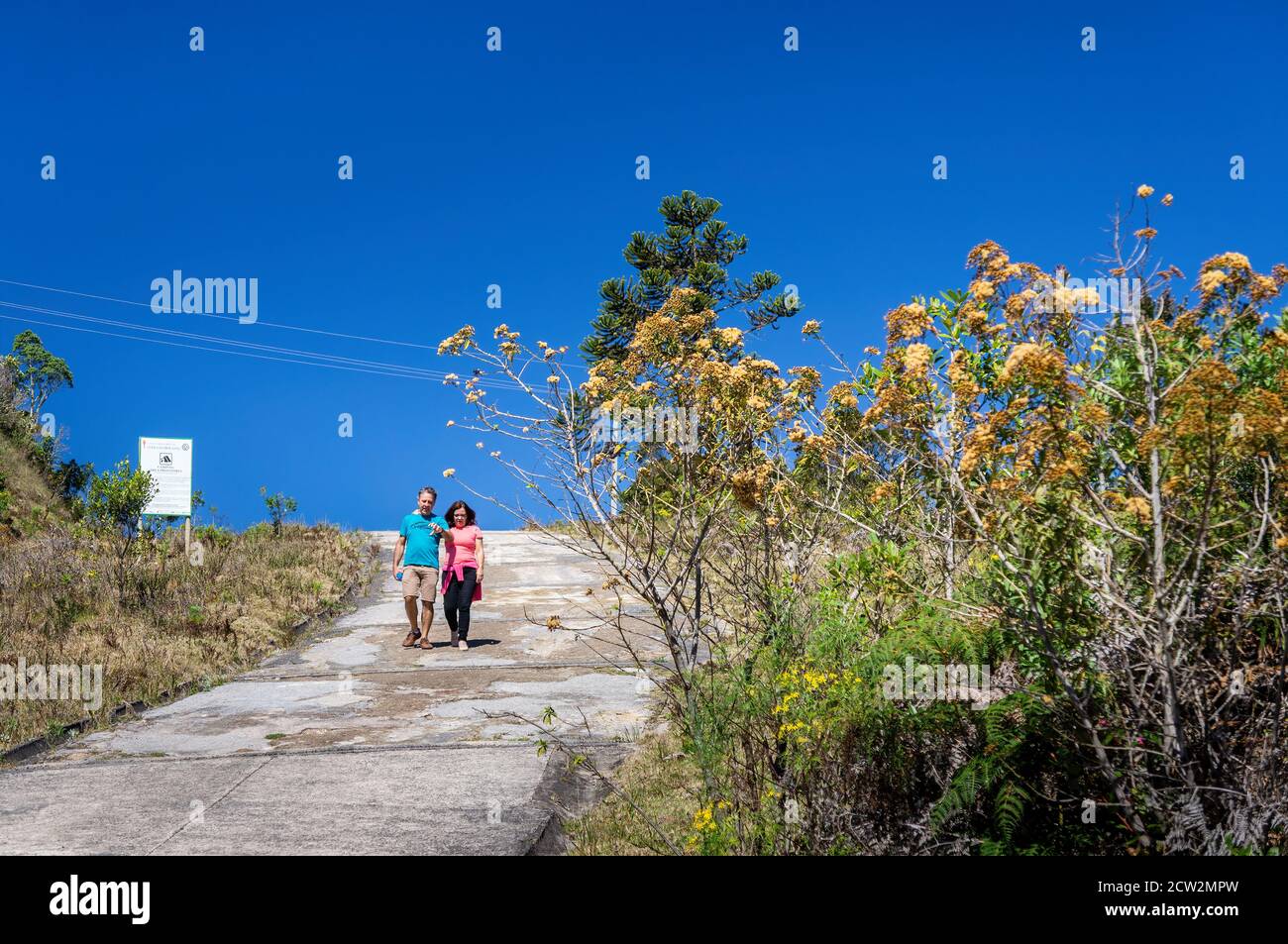 A couple walking down the rough path while take a conversation near the top of Pedra da Macela viewing spots and some bushes under sunny blue sky Stock Photo