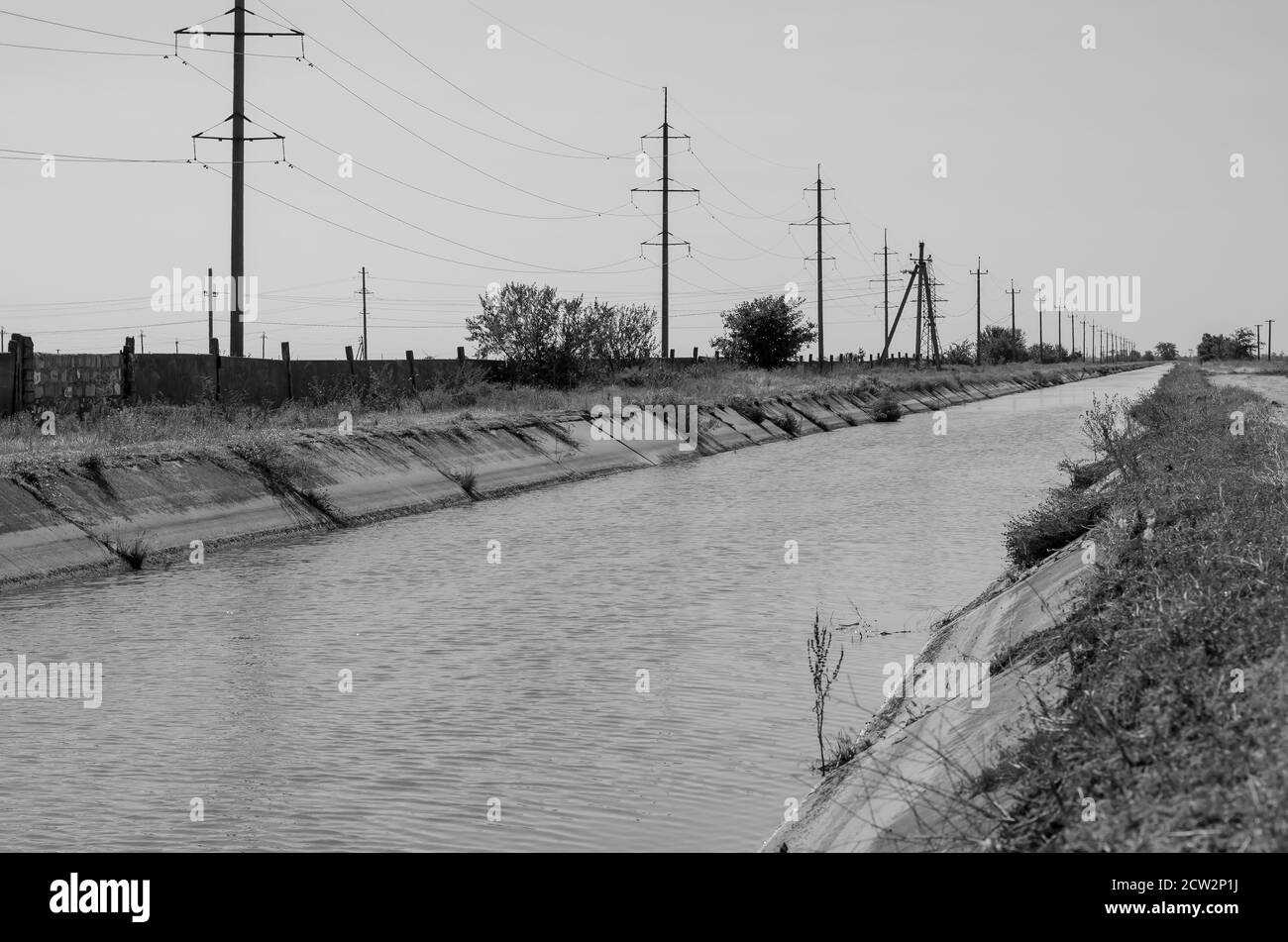 Irrigation agricultural channel with water. Power line along the canal for irrigation. Summer sunny day. Agribusiness. Monochrome Stock Photo