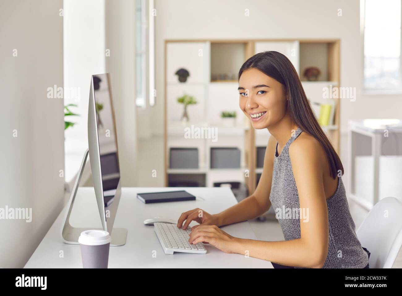 Young woman smiling and looking at camera while sitting at desk and working on computer Stock Photo