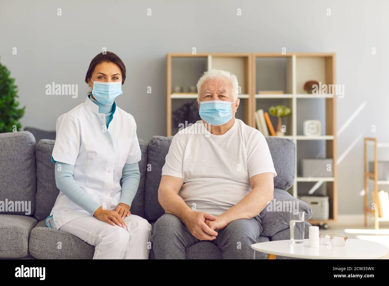Portrait of therapeutist and aged white-haired man, both wearing medical face masks, sitting on couch at home. Stock Photo