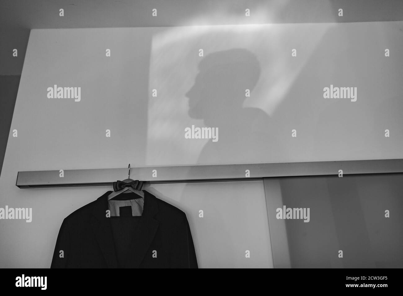 A man's jacket hangs on a hanger with a bow tie against the background of the shadow of a man, black and white photo. Stock Photo