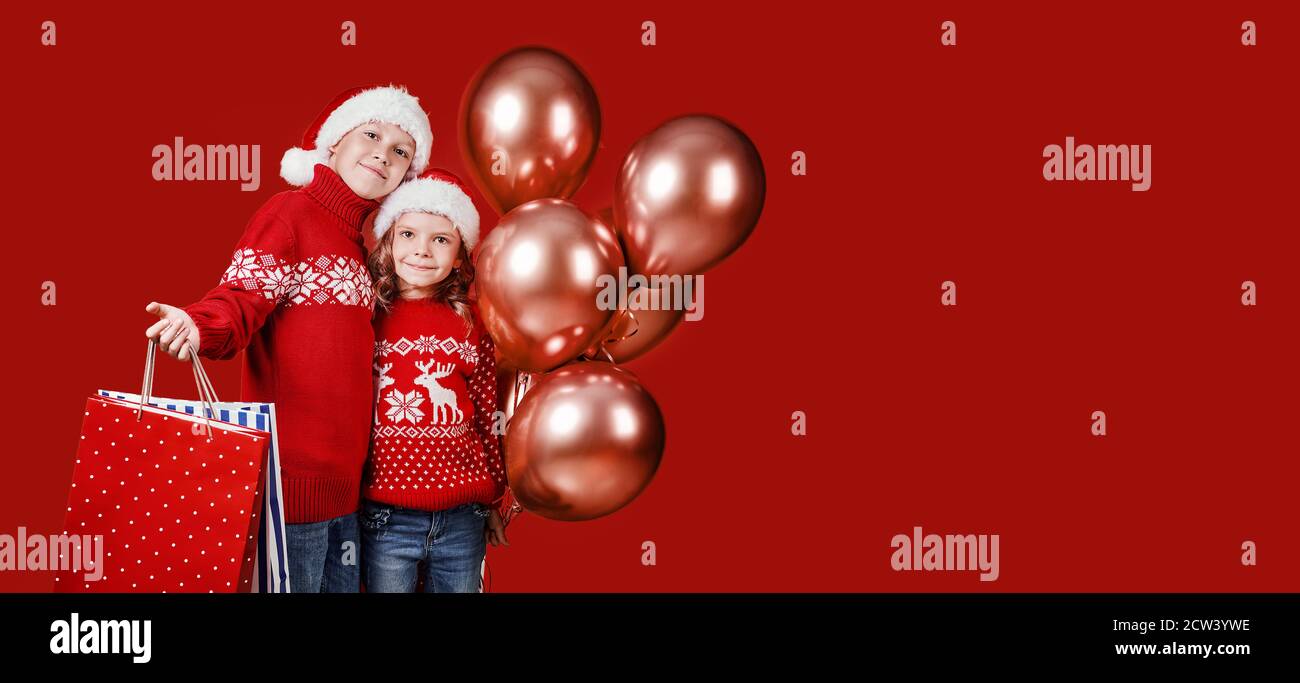 Cute children in red Santa hats, sweaters holding shopping bags and balloons on red background. Stock Photo