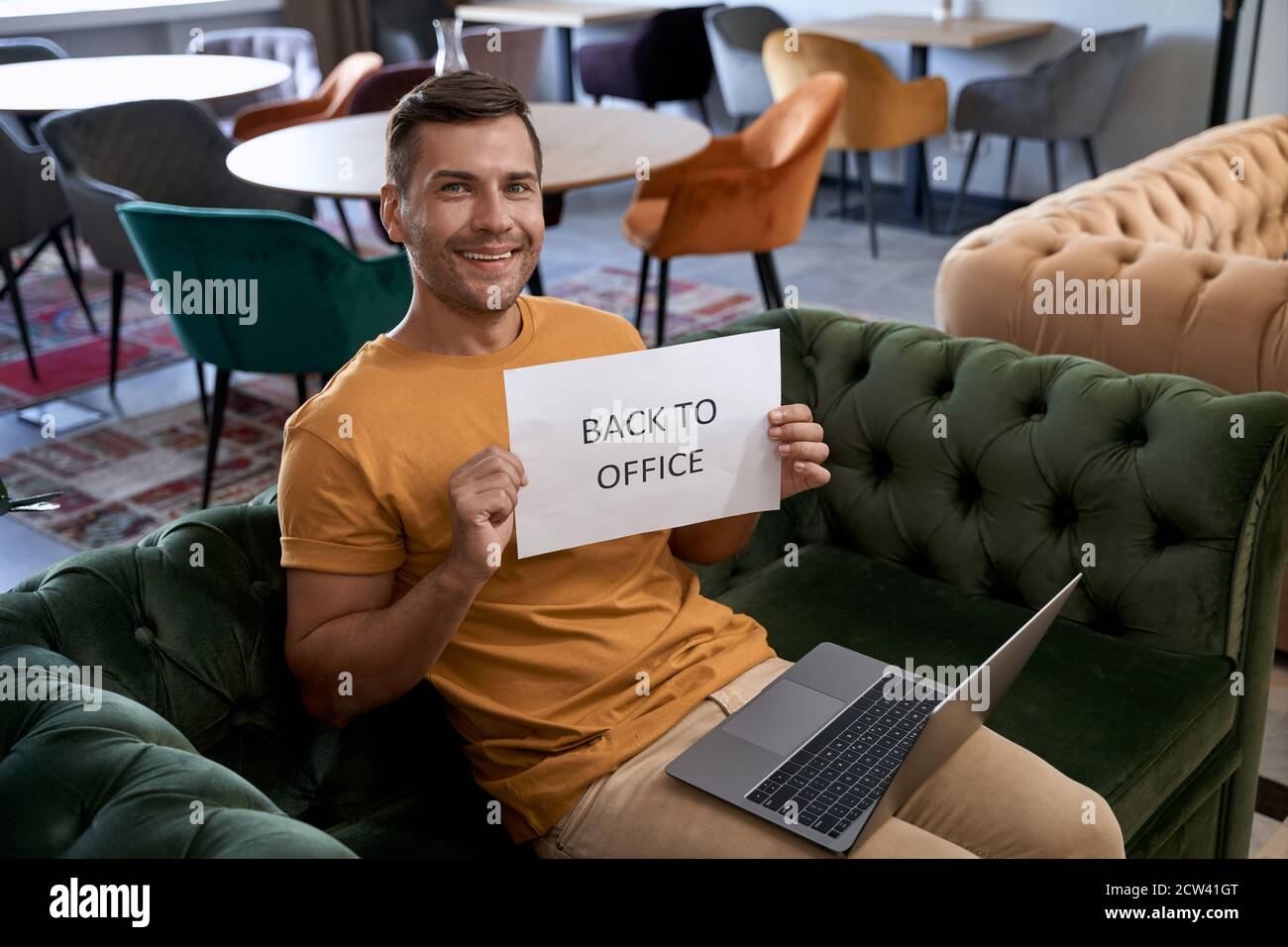 Young happy caucasian man showing paper with text BACK TO OFFICE and smiling at camera while working on laptop in the modern coworking space Stock Photo