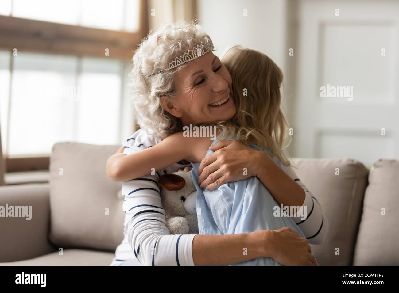 Affectionate grandmother wearing diadem on silver hair hugging little granddaughter Stock Photo