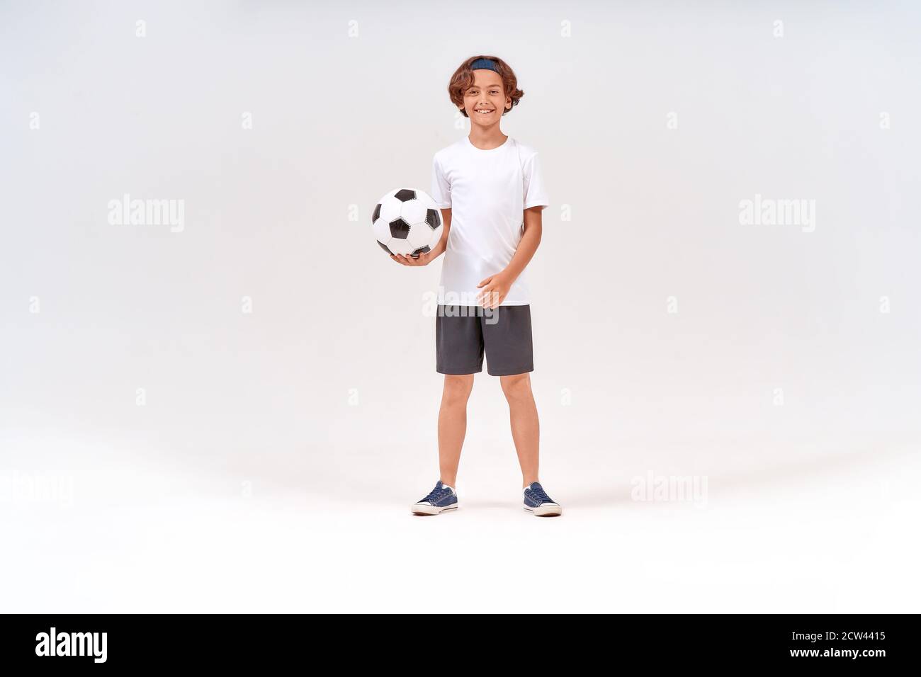 Being active. Full-length shot of a happy teenage boy with soccer ball looking at camera and smiling while standing isolated over grey background Stock Photo