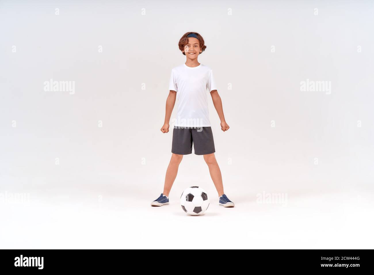 Full-length shot of a happy teenage boy playing football, looking at camera and smiling while standing isolated over grey background Stock Photo
