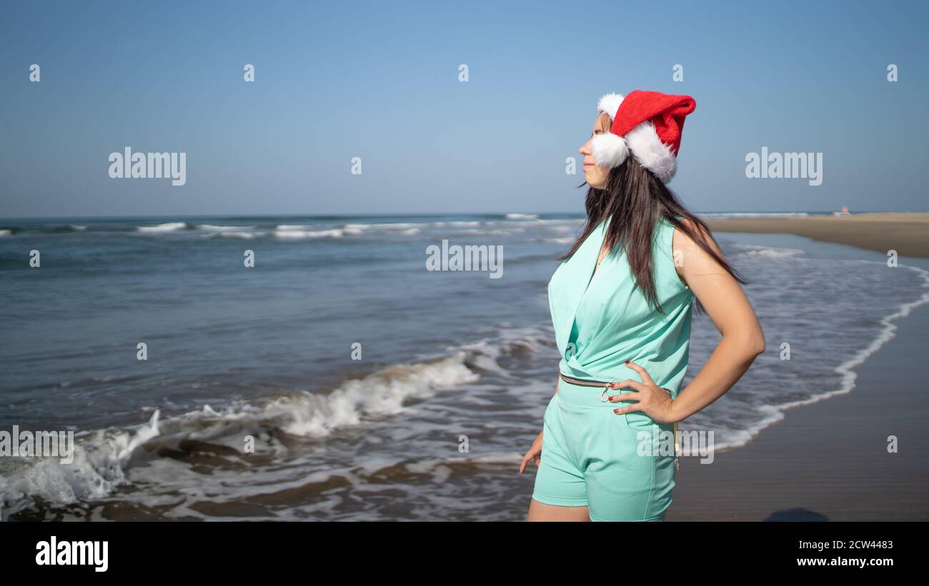 Young woman in Santa Claus hat standing on seashore in sunny weather ...
