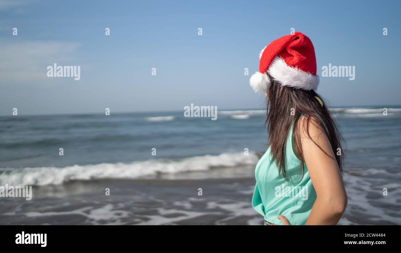 Young woman in Santa Claus hat standing on seashore in sunny weather ...