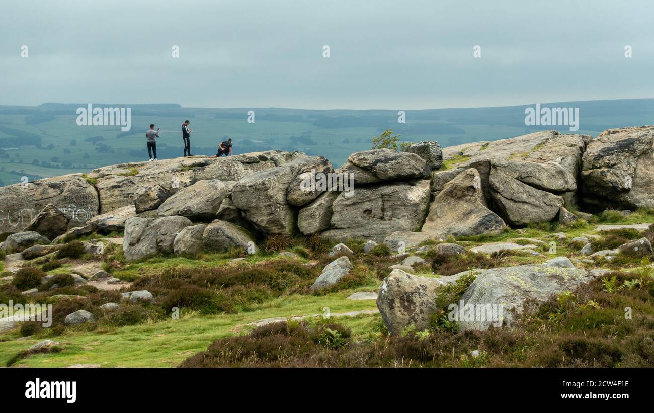 Three Asian lads enjoying the outdoors taking selfies & photos of each other - one doing a one-armed press up at the top of the Cow and Calf Rocks, UK Stock Photo