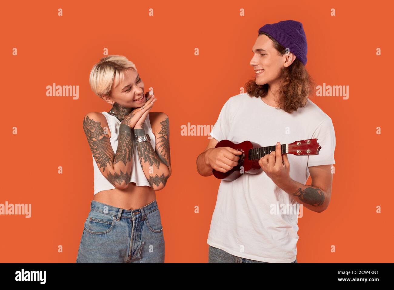 Freestyle. Young tattooed couple standing isolated on orange girlfriend looking at boyfriend touched while he playing ukulele smiling confident Stock Photo