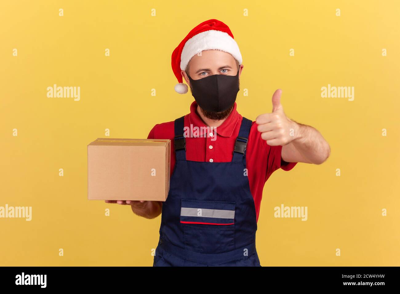 Logistic man in santa claus hat wearing medical protective mask and showing thumbs up holding cardboard box, satisfied with delivery service during qu Stock Photo