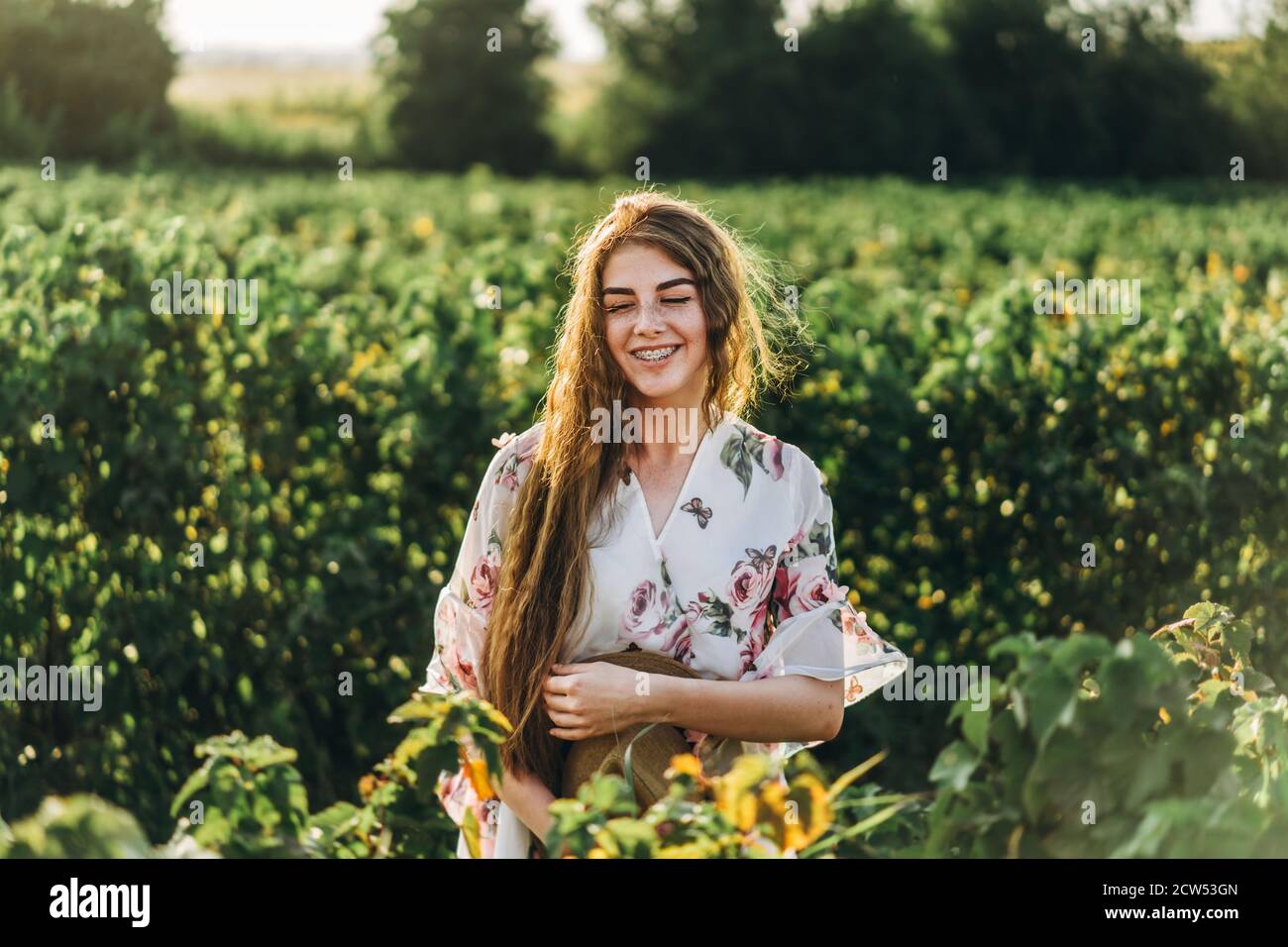 beautiful woman with long curly hair and freckles face on currant field background. Girl in a light dress walks in the summer sunny day. Stock Photo