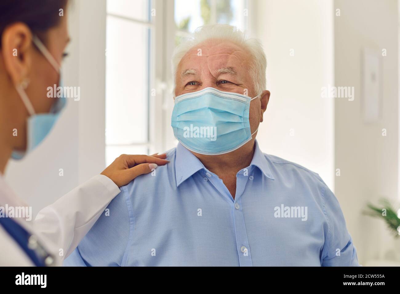 Happy senior white-haired patient in medical face mask trusting his young doctor Stock Photo