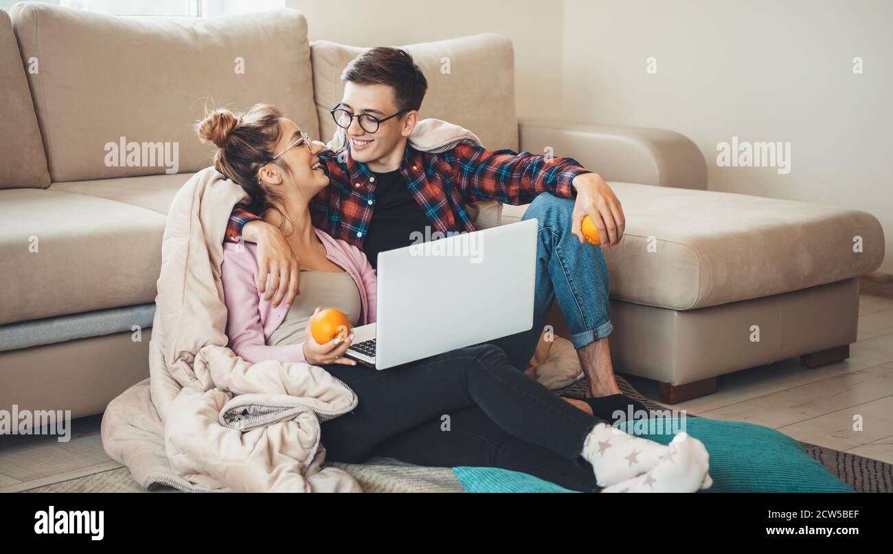 Cute smiling couple embracing on the floor near the sofa covered with a coverlet using a laptop and holding oranges Stock Photo