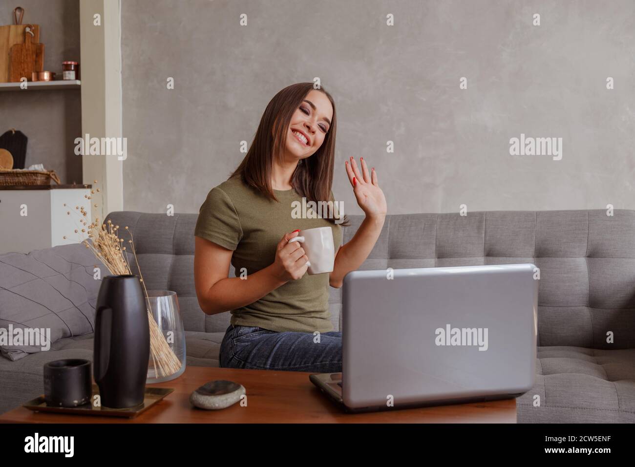 Portrait of pleasant young woman with mug looking at camera, waving hello. Pretty millennial lady communicating with parents via video call Stock Photo