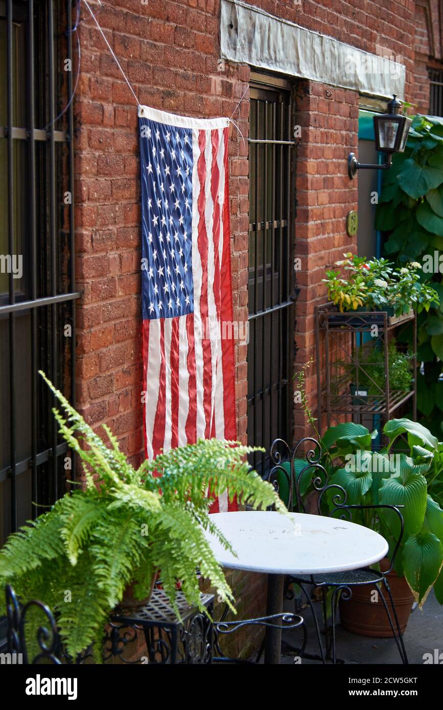 BROOKLYN, NY - SEPTEMBER 23 2020: An american Flag hangs on the side of an old brick house, in a small garden among plants and a wire table and chairs Stock Photo