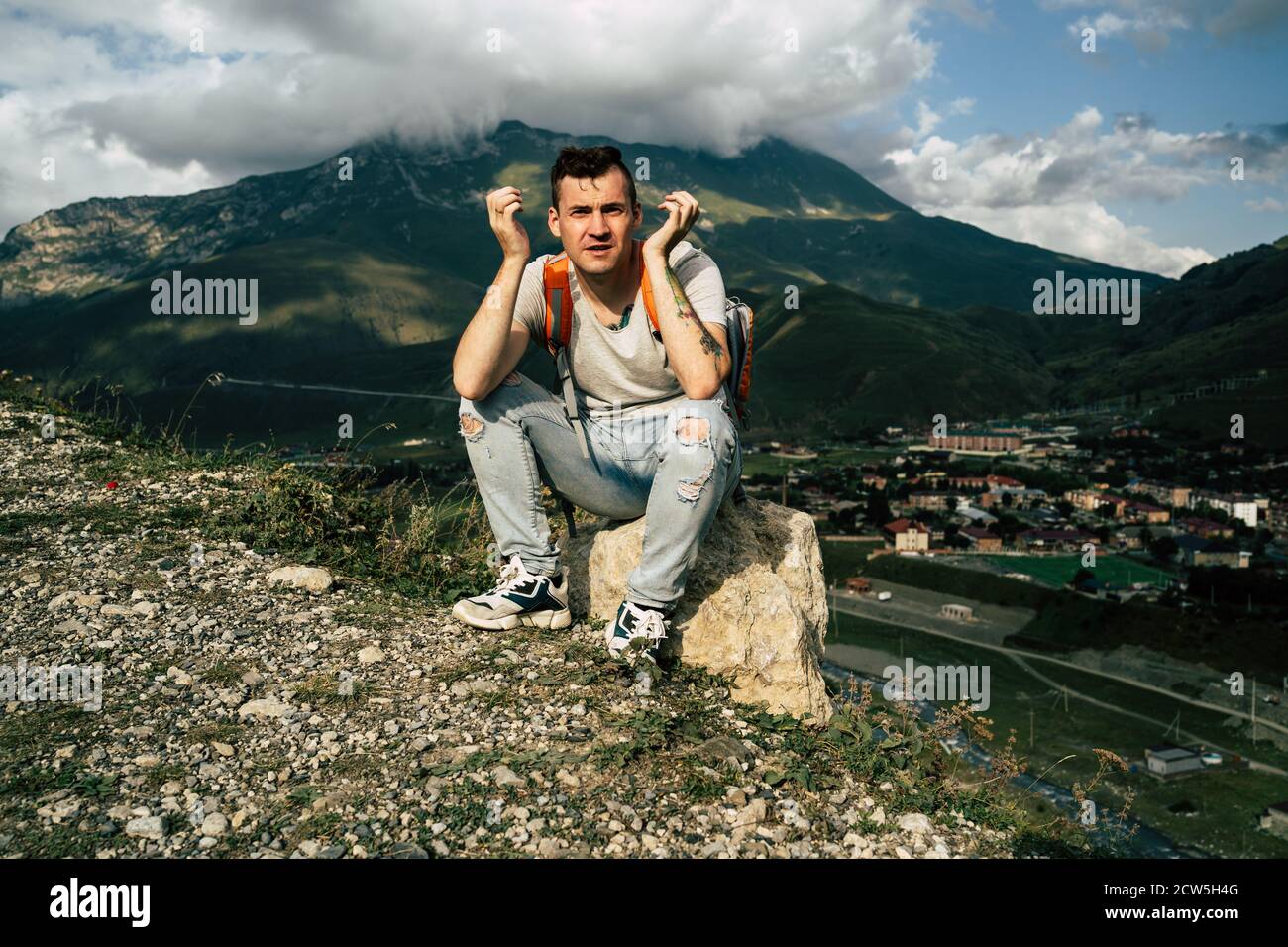 Young man grimaces, sitting on stone on background of village in mountains. Tired male tourist aping, resting on hill after active trekking in Stock Photo