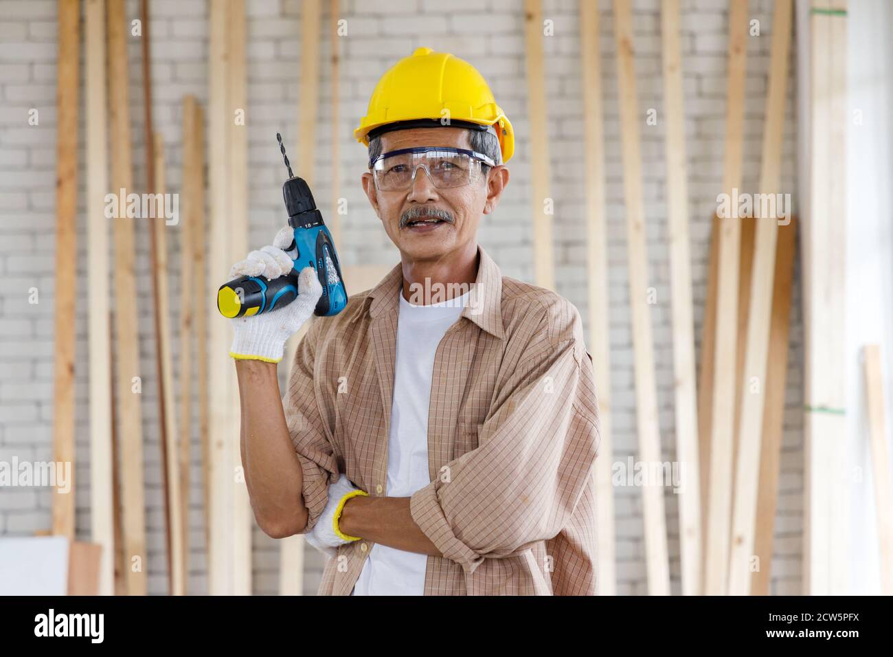 Asian senior man with hardhat and safety goggles hold electric drill in carpentry workshop. Concept senior people doing hobby after retirement. Stock Photo