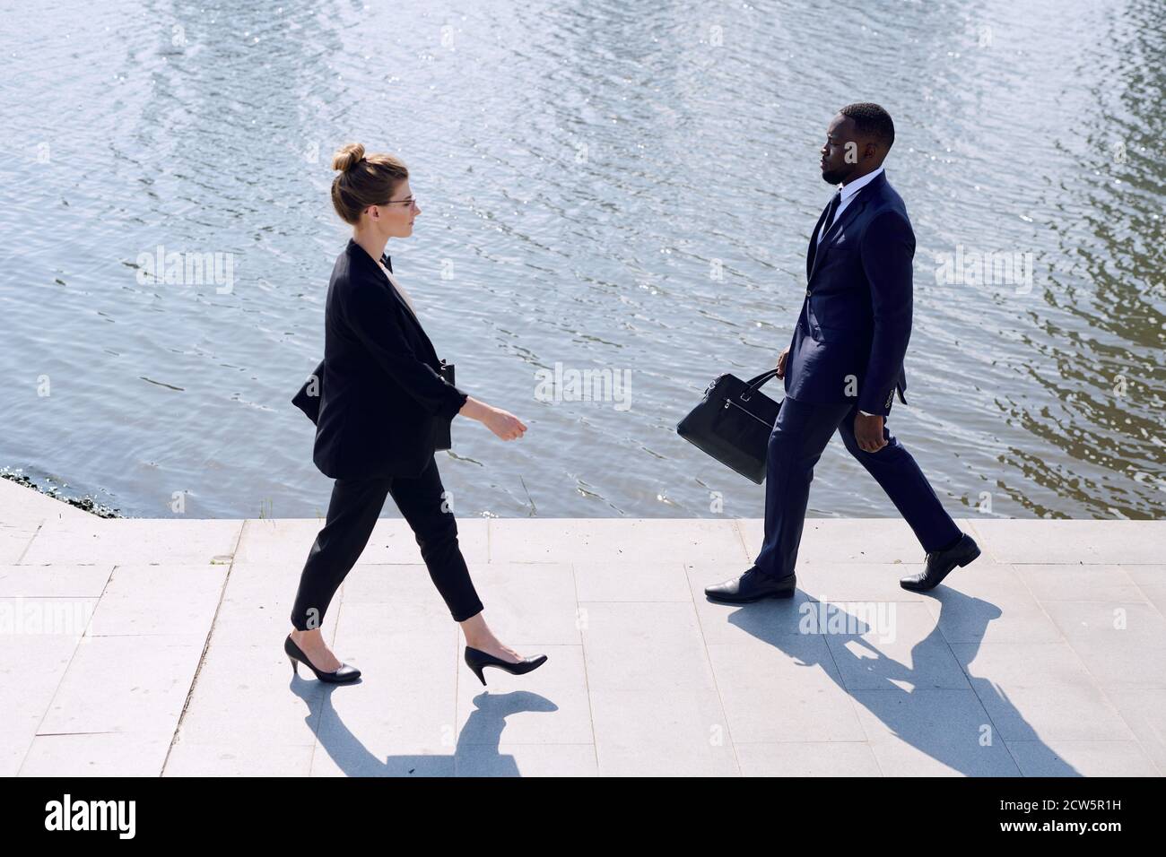 Young intercultural business partners in formalwear walking along riverside Stock Photo